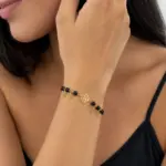 Pulsera dorada de acero con cuentas de ónix negro y dijes colgantes en forma de hoja y rosa, modelada en la muñeca.