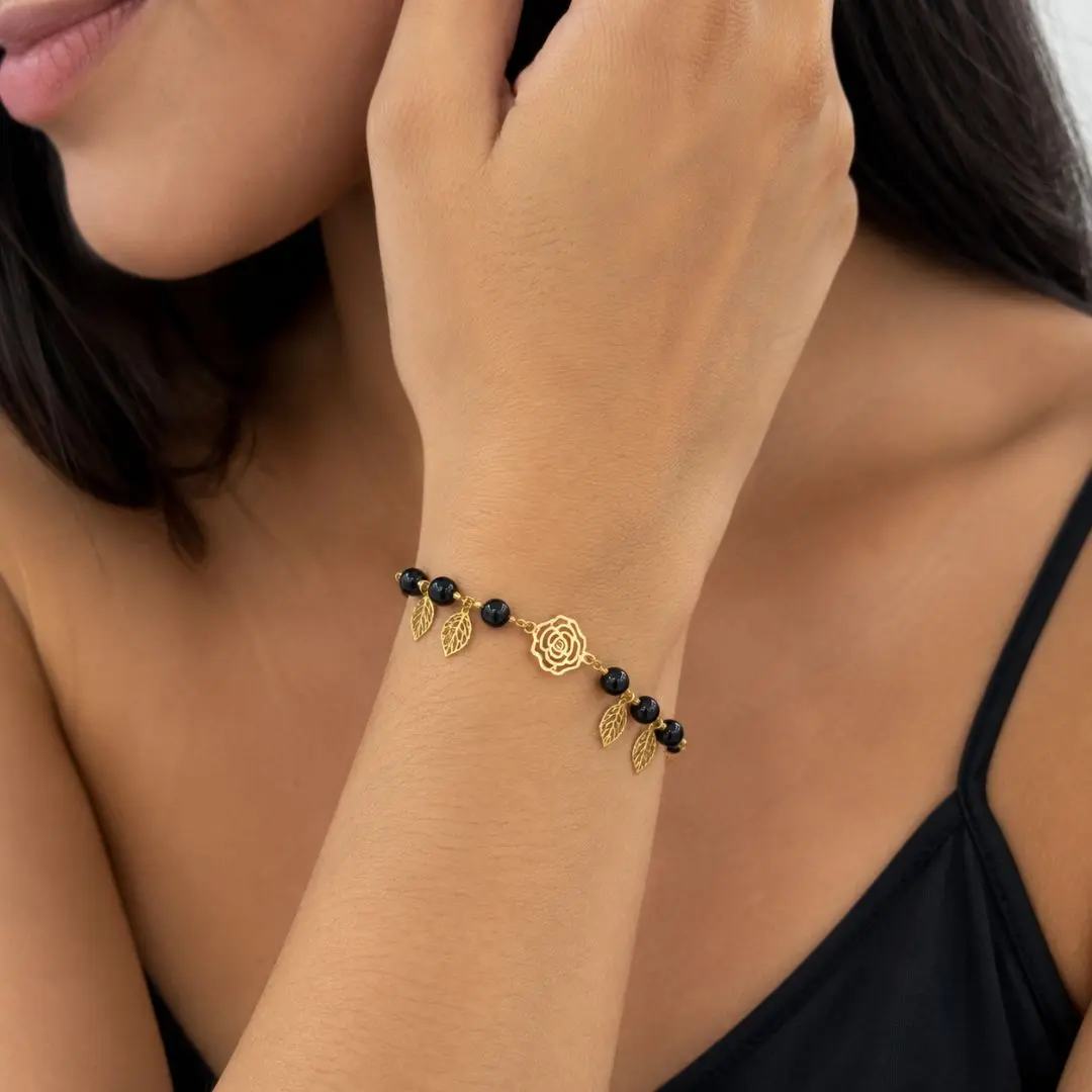 Pulsera dorada de acero con cuentas de ónix negro y dijes colgantes en forma de hoja y rosa, modelada en la muñeca.