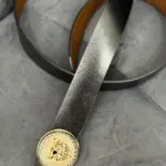 Correa de cuero negro texturizado para hombre con hebilla circular dorada con pedrería y emblema central, sobre fondo gris.