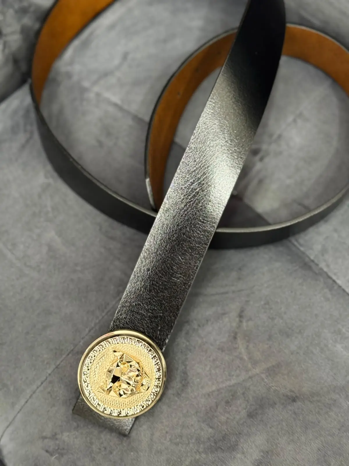 Correa de cuero negro texturizado para hombre con hebilla circular dorada con pedrería y emblema central, sobre fondo gris.