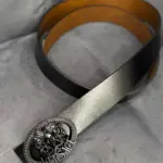 Correa de cuero negra con interior café y hebilla metálica ovalada con relieve de Medusa y la palabra Versace, sobre fondo gris texturizado.