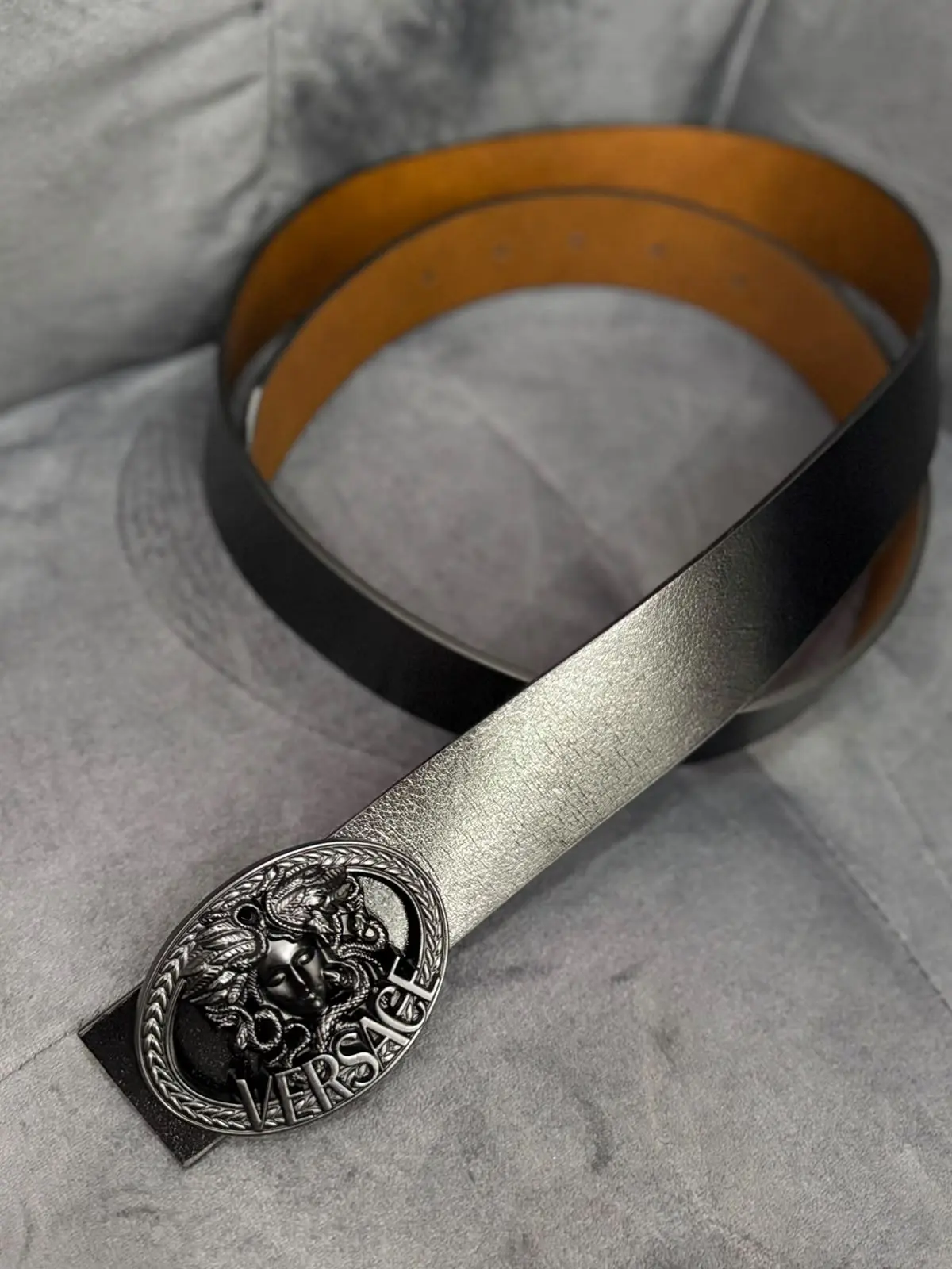 Correa de cuero negra con interior café y hebilla metálica ovalada con relieve de Medusa y la palabra Versace, sobre fondo gris texturizado.