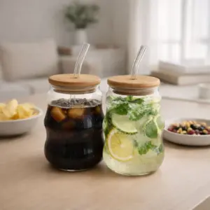 Dos vasos asimétricos de vidrio con tapa de madera y pitillo curvo, uno con bebida oscura y otro con limonada, sobre una mesa con snacks.