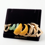 Cinco pares de candongas en tonos dorados, verde menta y amarillo, dispuestos sobre un soporte de terciopelo oscuro de Aura Jewels.