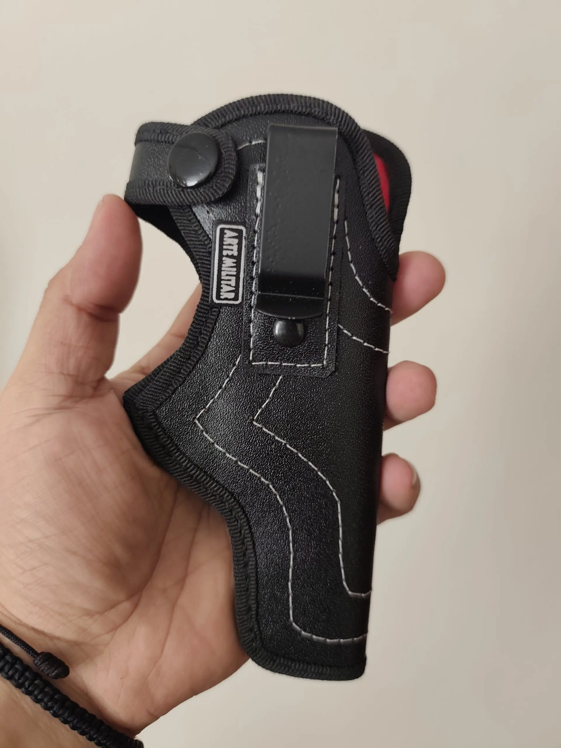 Funda negra texturizada de cuero ecológico para pistola, sostenida en una mano con gancho metálico y costuras blancas visibles.