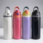 Cuatro termos deportivos de acero inoxidable con tapa abatible y asa, en colores blanco, rosa con condensación, lila con gotas y negro mate, alineados sobre superficie blanca.
