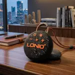 Parlante Bluetooth Lonio circular negro con textura granulada y detalles en naranja, con correa de transporte, sobre un escritorio de madera.