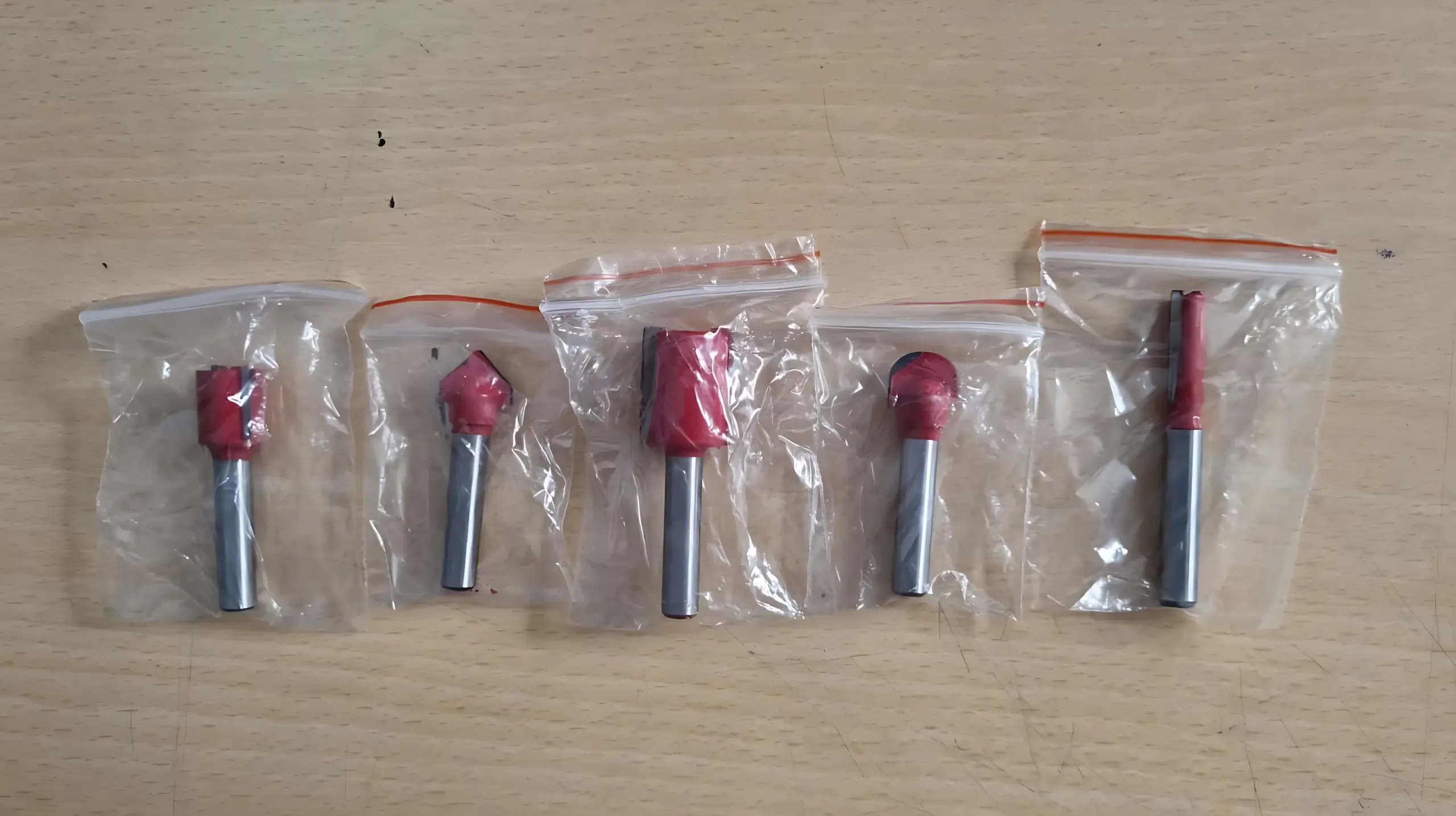 Cinco fresas para router con cuerpo rojo y vástago metálico, empacadas individualmente en bolsas plásticas transparentes sobre superficie de madera.