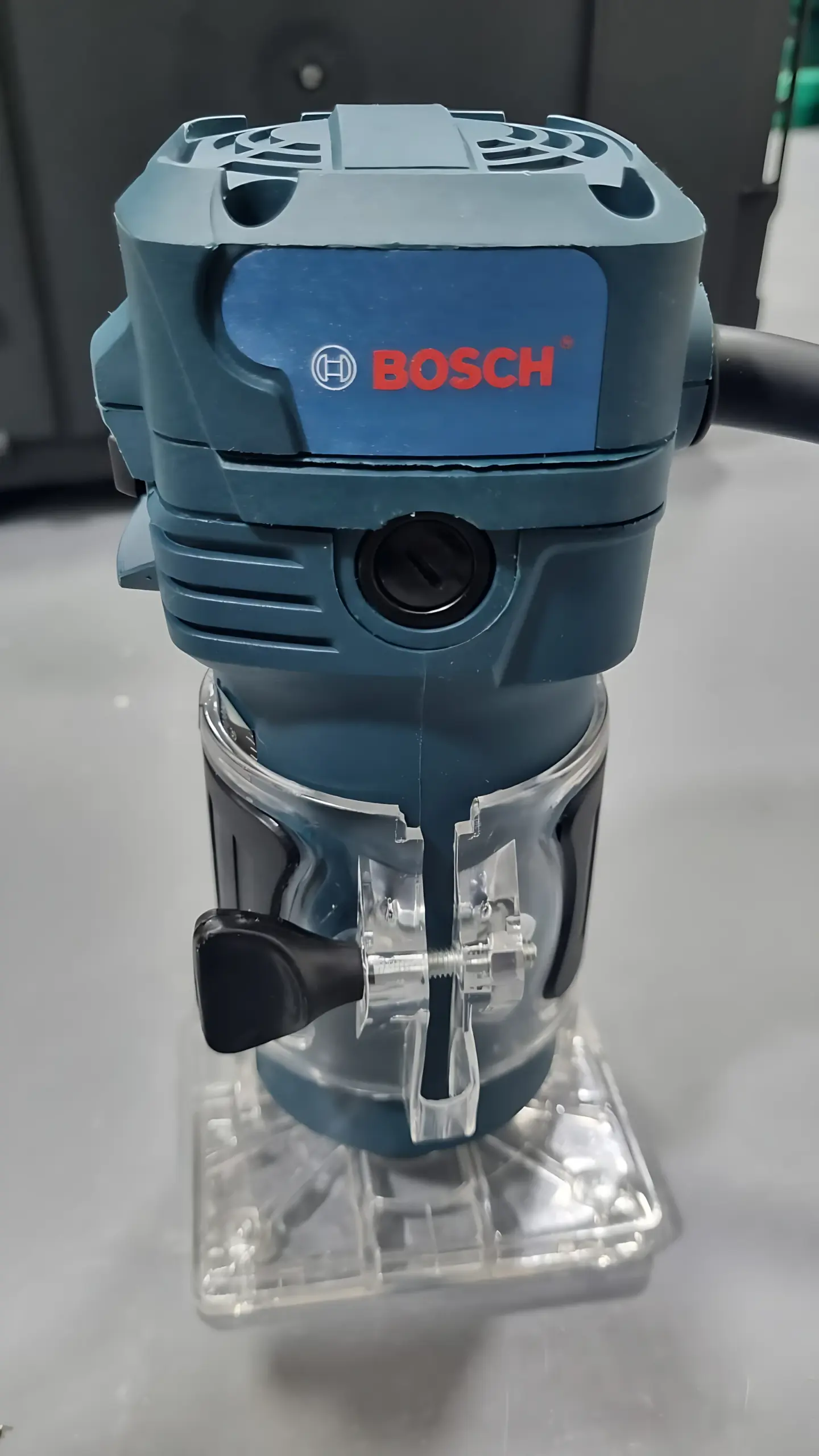 Rebordeadora eléctrica Bosch azul con base transparente fija, perilla de ajuste negro y mango lateral con cable, vista frontal sobre superficie gris.