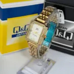 Reloj Q&Q cuadrado dorado para dama con pedrería en el bisel, pulso metálico, sobre soporte transparente y cajas de la marca.