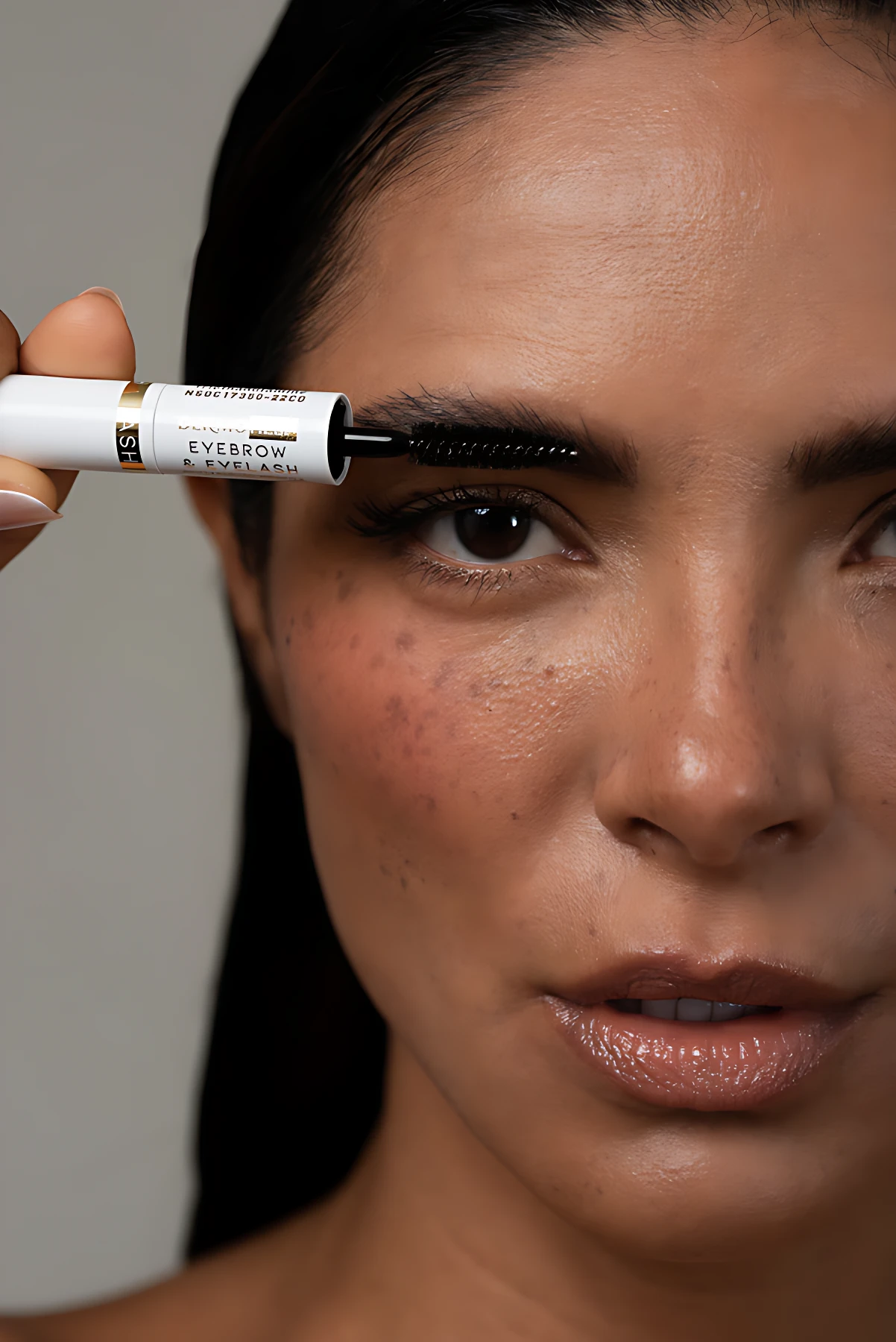 Mujer aplicando tratamiento para cejas con aplicador de cepillo negro desde un envase blanco con detalles dorados.