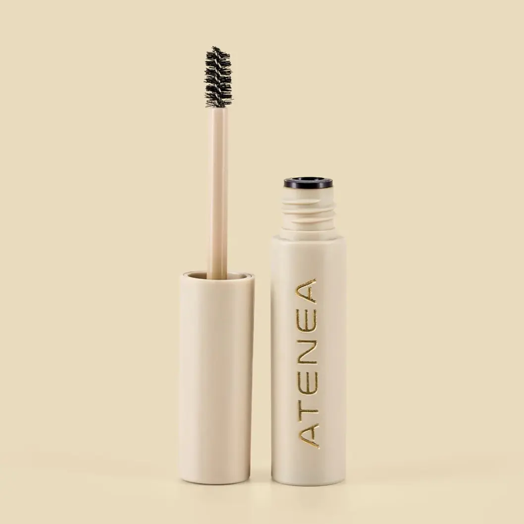Gel fijador de cejas transparente Atenea en envase beige con letras doradas y aplicador de cepillo negro, presentación tamaño viaje.