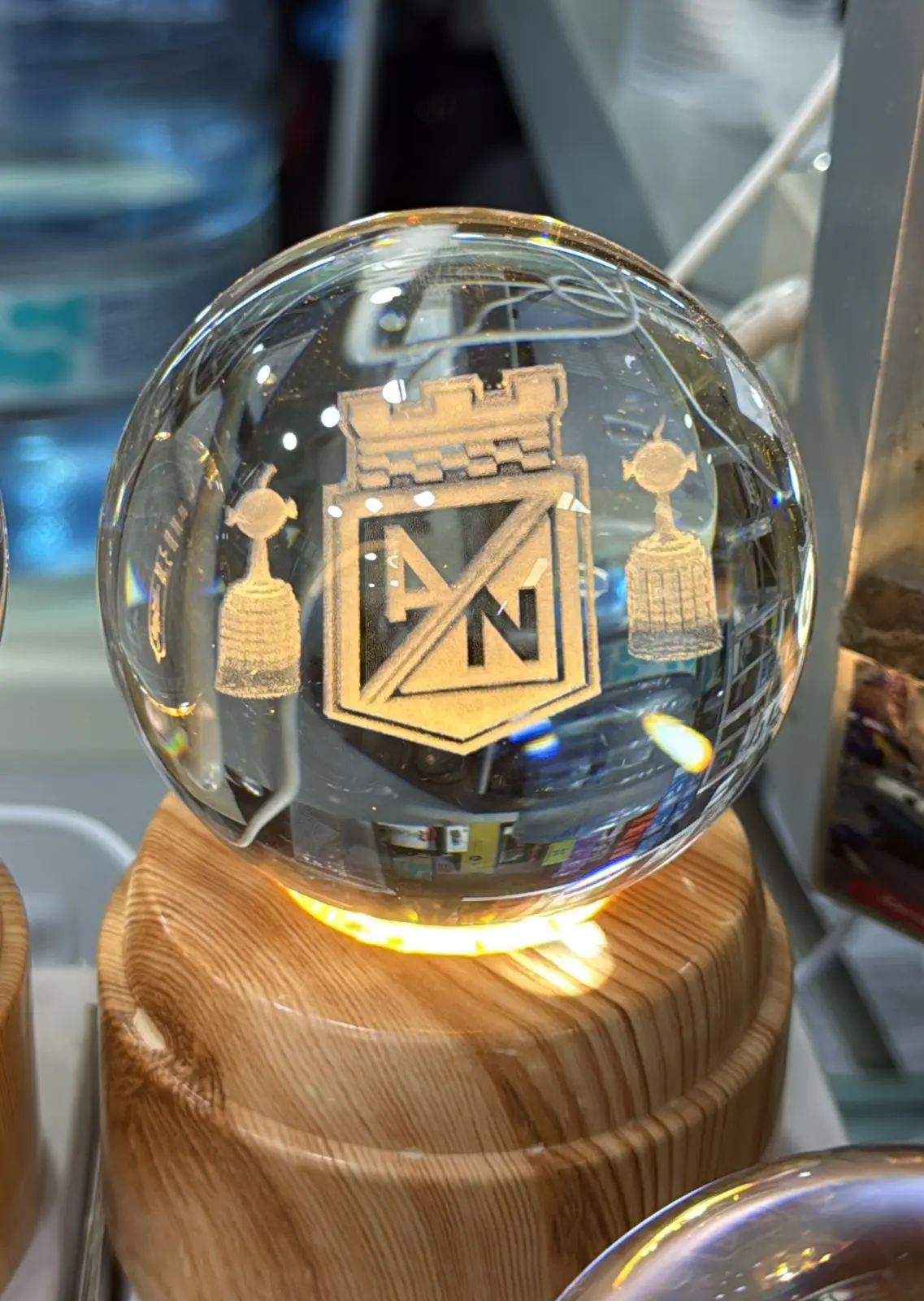Lámpara LED esférica de cristal con grabado láser del escudo de Atlético Nacional y dos copas, sobre base de madera iluminada con luz cálida.