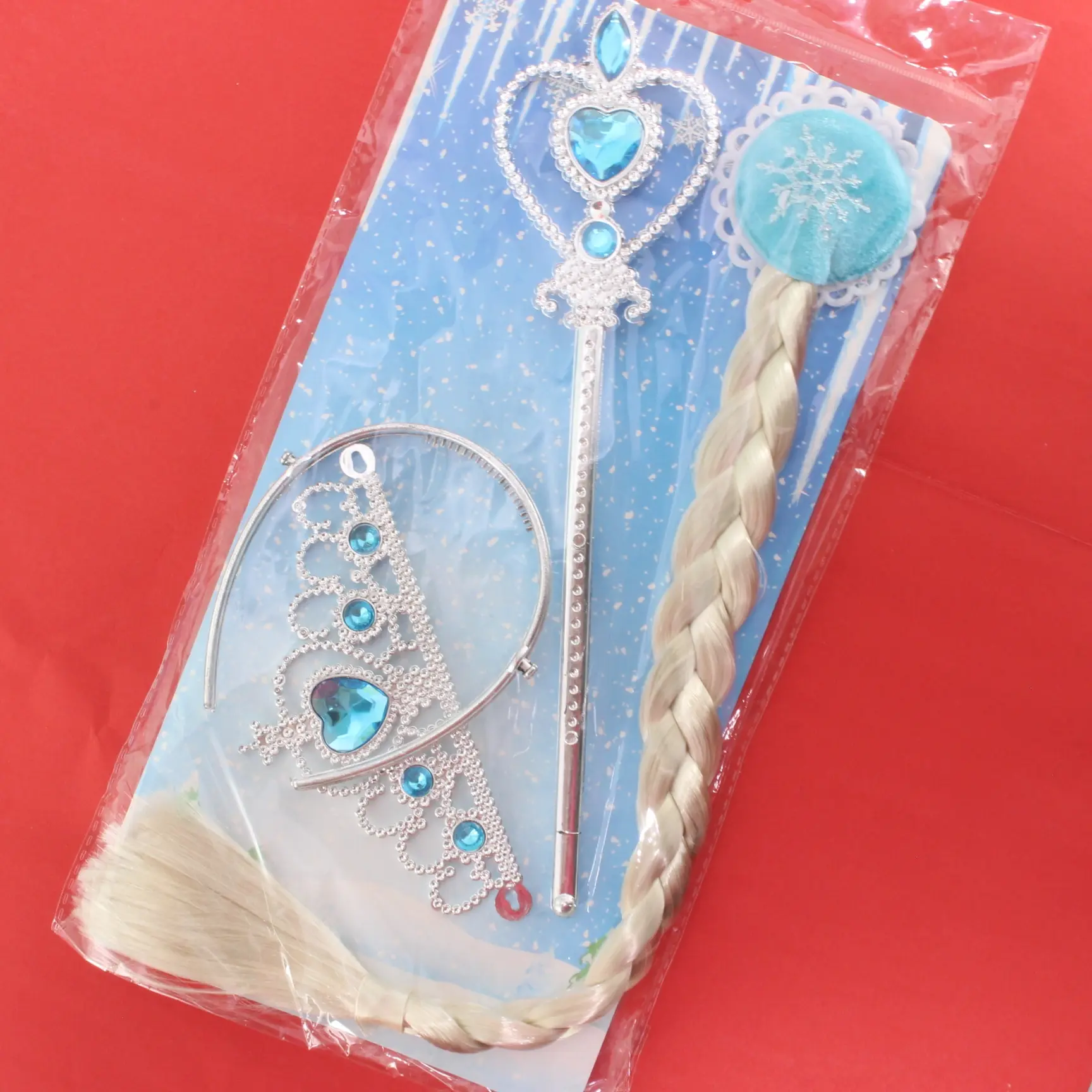 Kit de disfraz Frozen en blister sobre fondo rojo; incluye cetro plateado con gema azul, diadema brillante y trenza rubia larga.