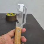 Pelador dentado de acero inoxidable con mango de madera clara sujeto en mano sobre mesa oscura y cinta adhesiva al fondo.