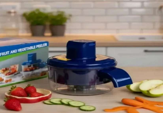 Pelador eléctrico de frutas y verduras inalámbrico en color azul oscuro, sobre una mesa de madera con rodajas de pepino, fresas y manzanas.
