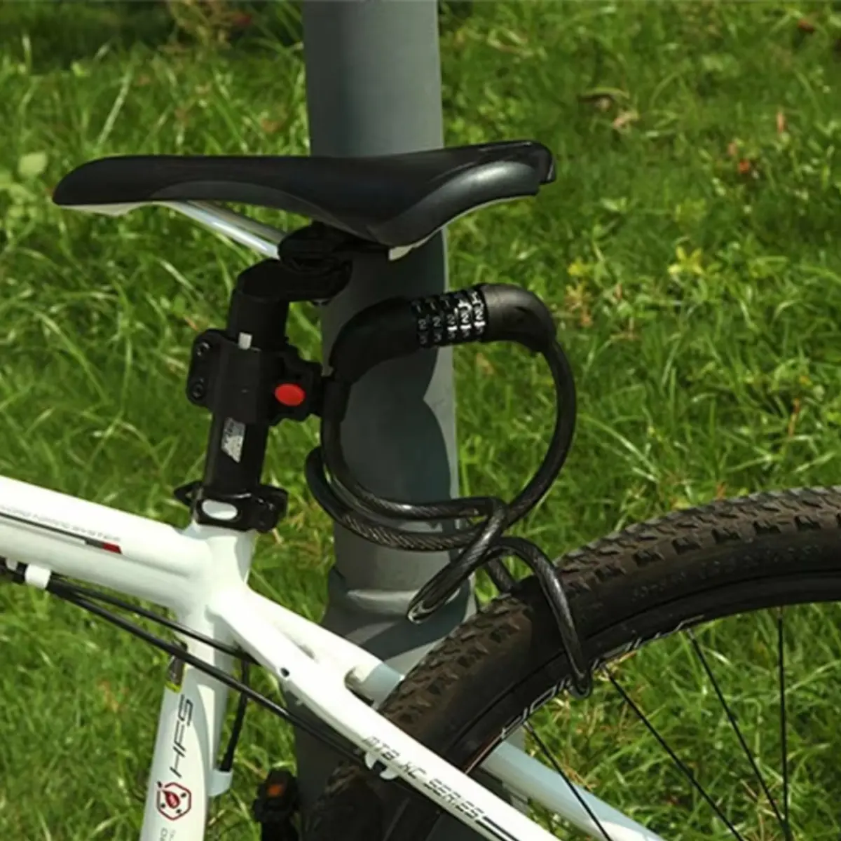 Candado guaya de acero negro con clave numérica, asegurando el sillín de una bicicleta blanca a un poste metálico sobre césped.