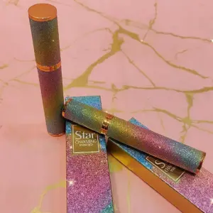 Máscara de pestañas con tubo texturizado brillante en tonos tornasol verde y morado, sobre cajas decoradas en superficie rosa con vetas doradas.