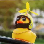 Pato decorativo amarillo con casco verde de hélice, gafas oscuras y cadena dorada, sobre el tablero de un vehículo con fondo verde desenfocado.