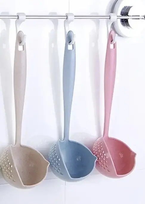Tres cucharones plásticos de trigo con colador integrado, en colores beige, azul y rosa, colgados de ganchos en barra de pared.