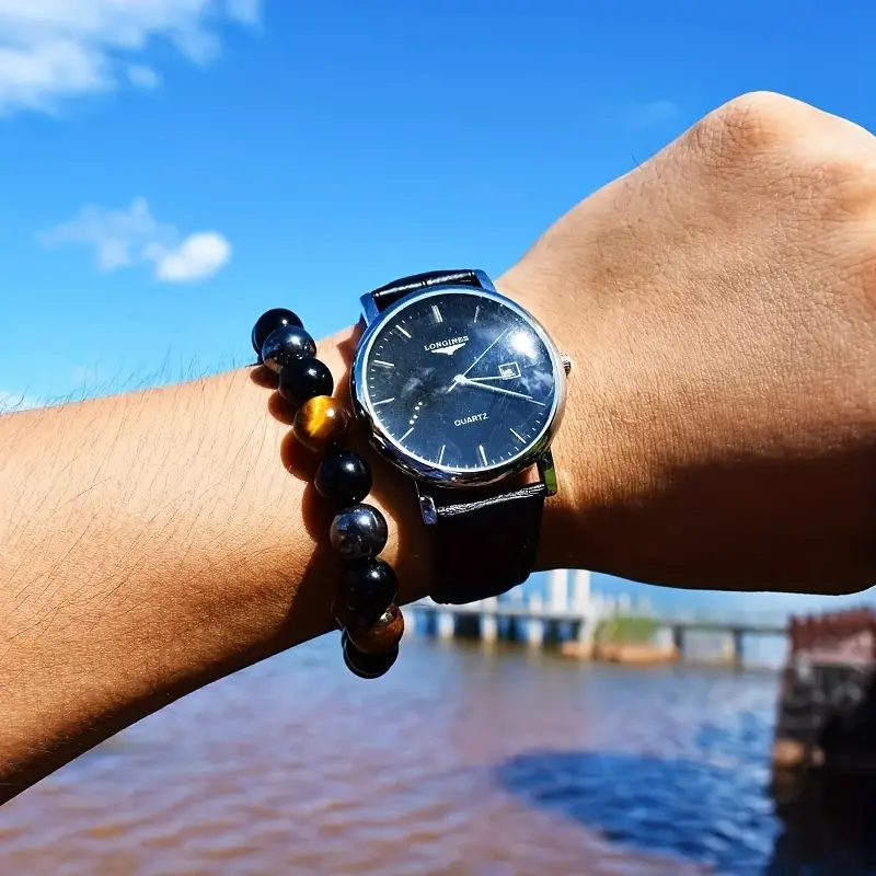 Muñeca con reloj Longines de esfera negra y correa de cuero, junto a una pulsera de protección con cuentas de hematita, obsidiana y ojo de tigre.