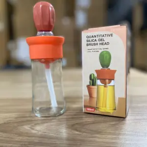 Botella dispensadora de aceite de 180 ml con tapa y gotero de silicona rosada, junto a su caja de empaque sobre superficie de madera.