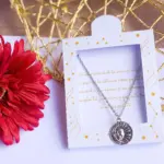 Collar plateado con dije de sol y luna en caja blanca decorada con estrellas y texto romántico, al lado de una flor roja.