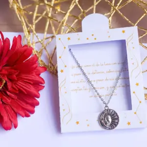 Collar plateado con dije de sol y luna en caja blanca decorada con estrellas y texto romántico, al lado de una flor roja.
