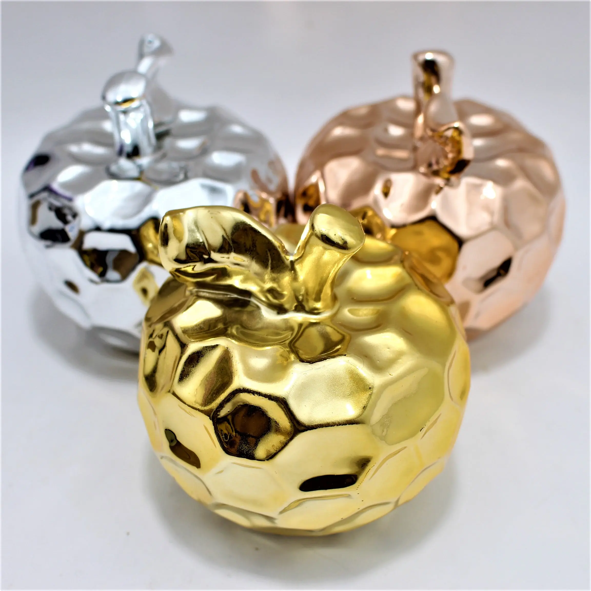 Manzanas decorativas de cerámica brillante en colores dorado, plateado y bronce, con textura facetada sobre fondo blanco.