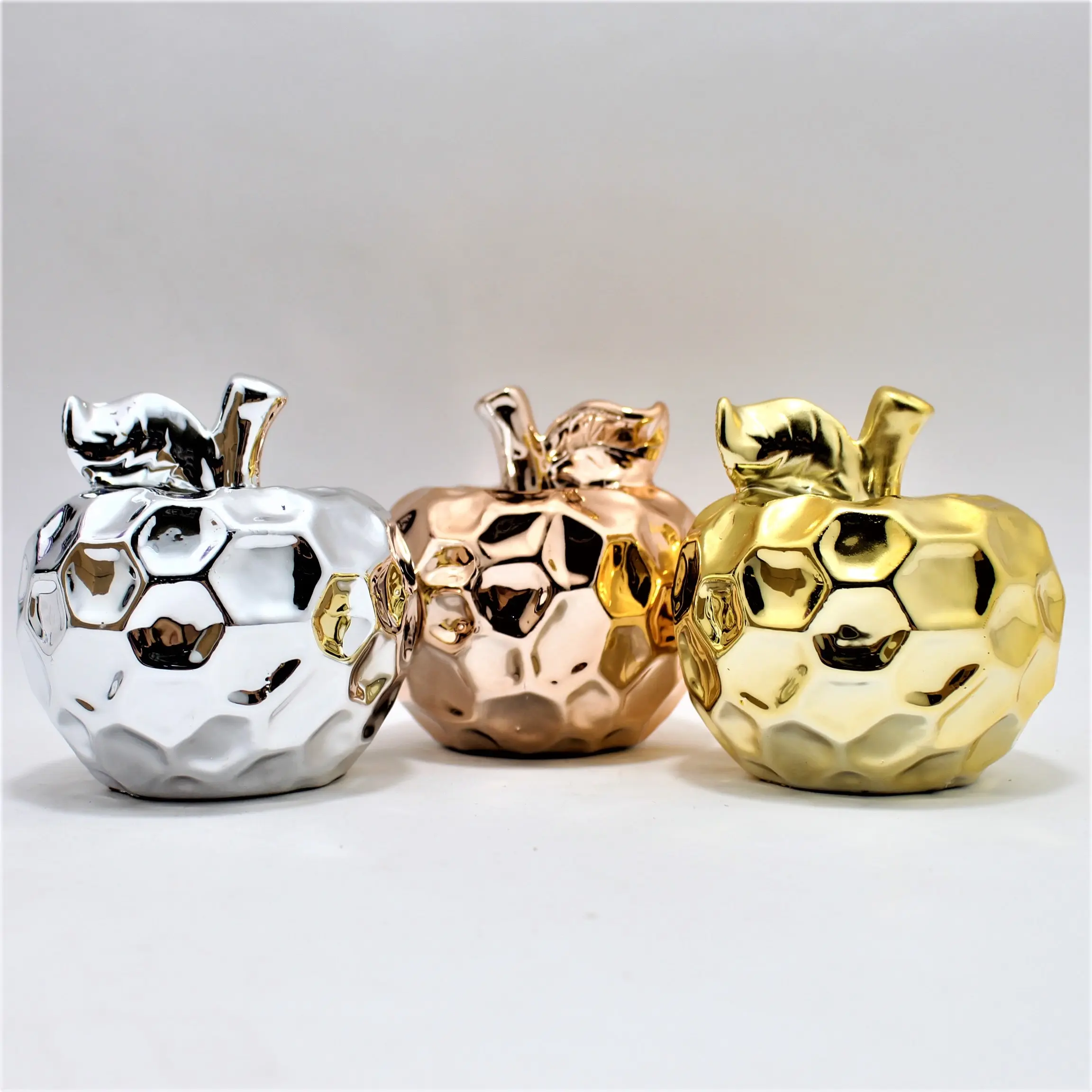 Tres manzanas decorativas de cerámica brillante con acabado facetado en colores plata, cobre y oro, alineadas sobre una superficie blanca.