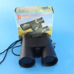 Binoculares negros de 6X35mm en primer plano junto a su caja de cartón con diseño de camuflaje, sobre fondo azul claro.
