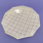 Plato decagonal de 23 cm en color blanco con diseño de cuadrícula en tono oro rosa, envuelto en plástico transparente sobre fondo morado.