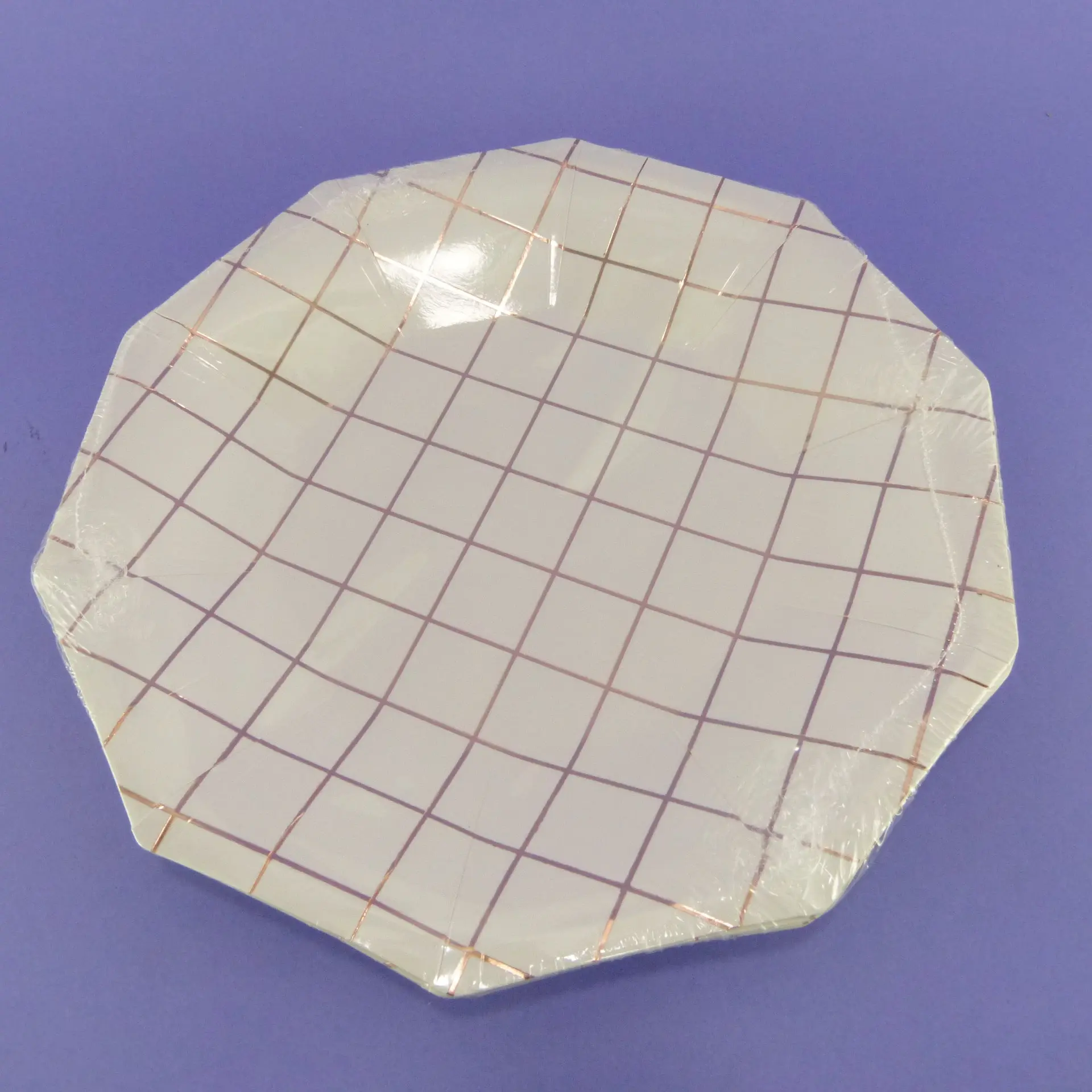 Plato decagonal de 23 cm en color blanco con diseño de cuadrícula en tono oro rosa, envuelto en plástico transparente sobre fondo morado.