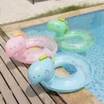 Set x3 flotadores inflables de dinosaurio con escarcha para piscina, en colores rosado, verde y azul, sobre muelle de madera junto al agua.