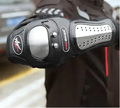 Codera de protección negra para motociclista con carcasa articulada de acero inoxidable plateado y dos remaches metálicos frontales visible en primer plano.