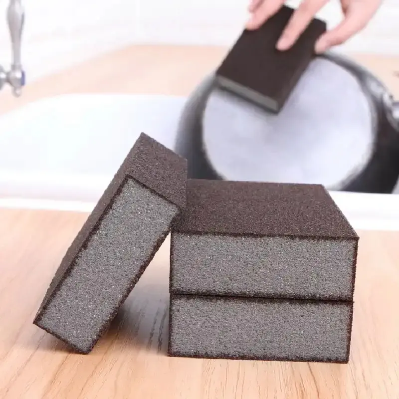 Esponjas Ninja de textura abrasiva color café y espuma gris, dispuestas en grupo sobre una mesa de madera, una de ellas limpiando el fondo de una olla.