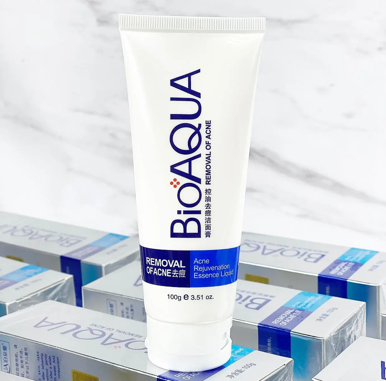 Tubo blanco de crema limpiadora Bioaqua 100g para remover acné y controlar grasa, de pie sobre cajas del producto.