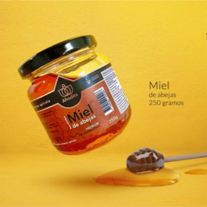 Tarro inclinado de miel de abejas de 250 gramos sobre fondo amarillo, con etiqueta marrón, cuchara de madera y charco de miel fresca.