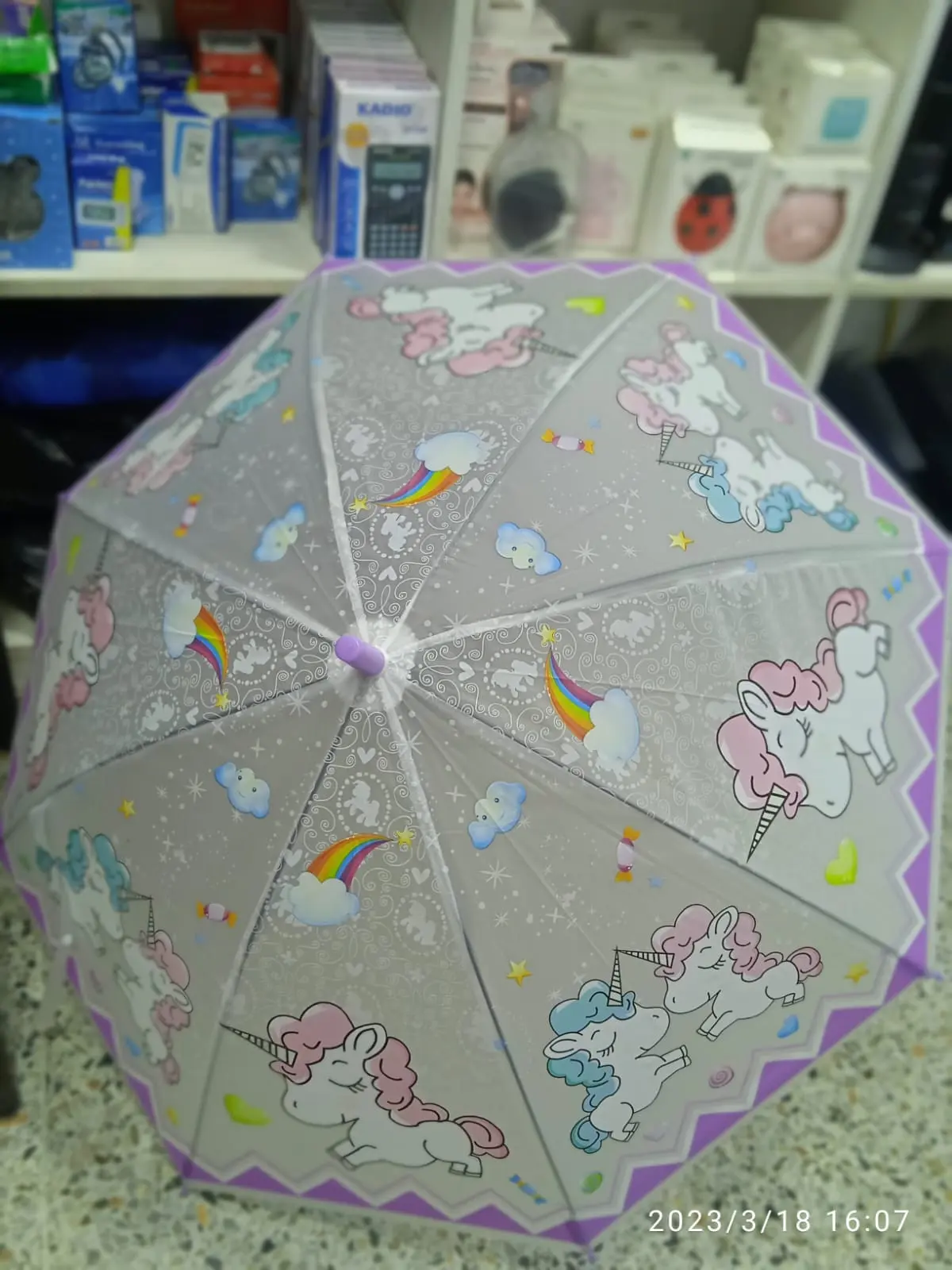 Sombrilla infantil gris con estampado de unicornios, arcoíris y nubes, borde morado y estructura transparente.