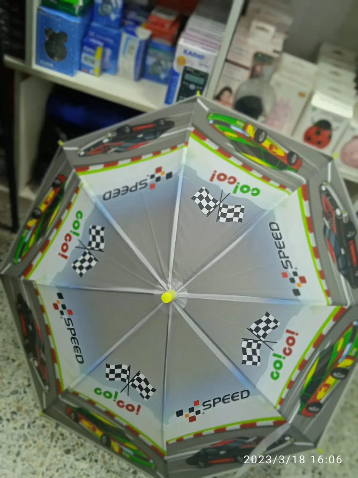 Sombrilla infantil con diseño de carreras, carros, banderas a cuadros y texto SPEED en colores gris, blanco y verde. Vista desde arriba.