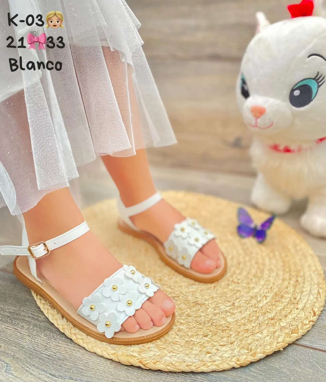 Sandalia plana infantil blanca con tira ajustable y flores decorativas sobre suela flexible, puesta en un pie de niña.