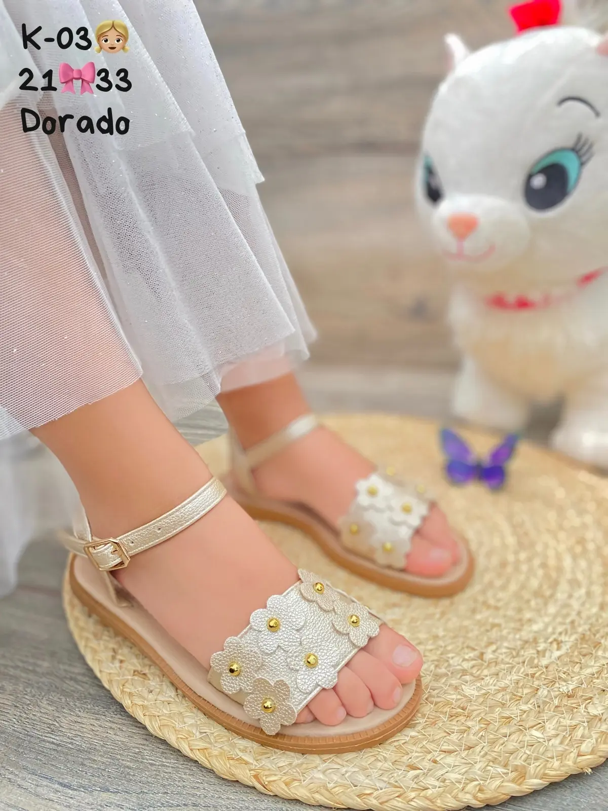 Sandalia plana infantil dorada con tira de flores decorativas y hebilla al tobillo, sobre una esterilla circular con fondo de madera.