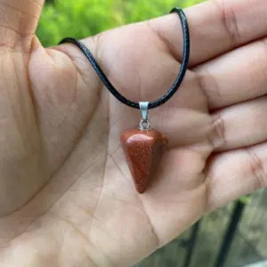 Collar péndulo de piedra arenisca marrón brillante y diamantada, en forma de cono, suspendido en una cuerda negra sobre la palma de una mano.