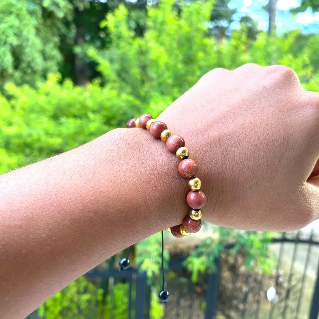 Pulsera de piedra natural café rojizo y cuentas doradas en la muñeca, con cordón negro y hematita, sobre fondo de vegetación verde desenfocado.