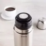 Termo bala de acero inoxidable plateado con dispensador negro y botón blanco, junto a una taza metálica. Al fondo, una taza con café.