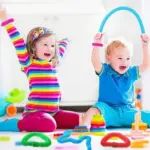 Niña y niño felices jugando con tubos elásticos sensoriales de colores, apilables y piezas de construcción sobre una superficie blanca.