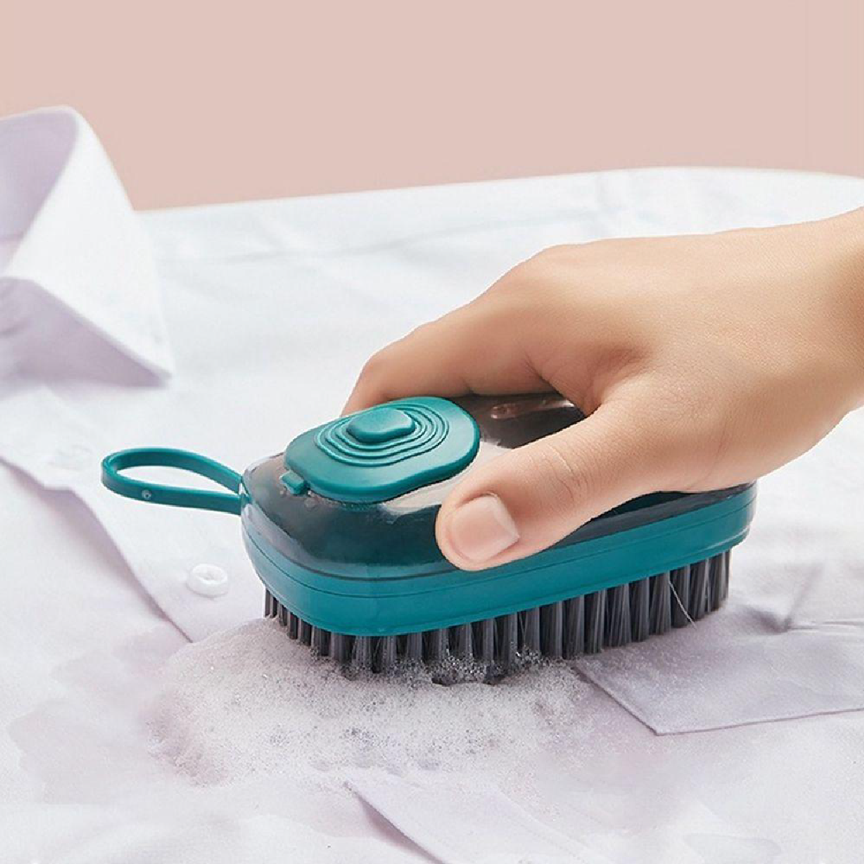 Cepillo dispensador de detergente color turquesa con cerdas negras limpiando una mancha con espuma sobre una camisa blanca.