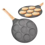 Sartén antiadherente gris con siete cavidades circulares, mango de madera y acabado moteado, mostrada vacía y con panqueques cocinándose en su interior.