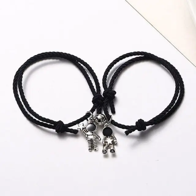 Par de pulseras de cordón negro ajustable con dijes de astronautas plateados y detalles negros, dispuestas juntas sobre fondo blanco.