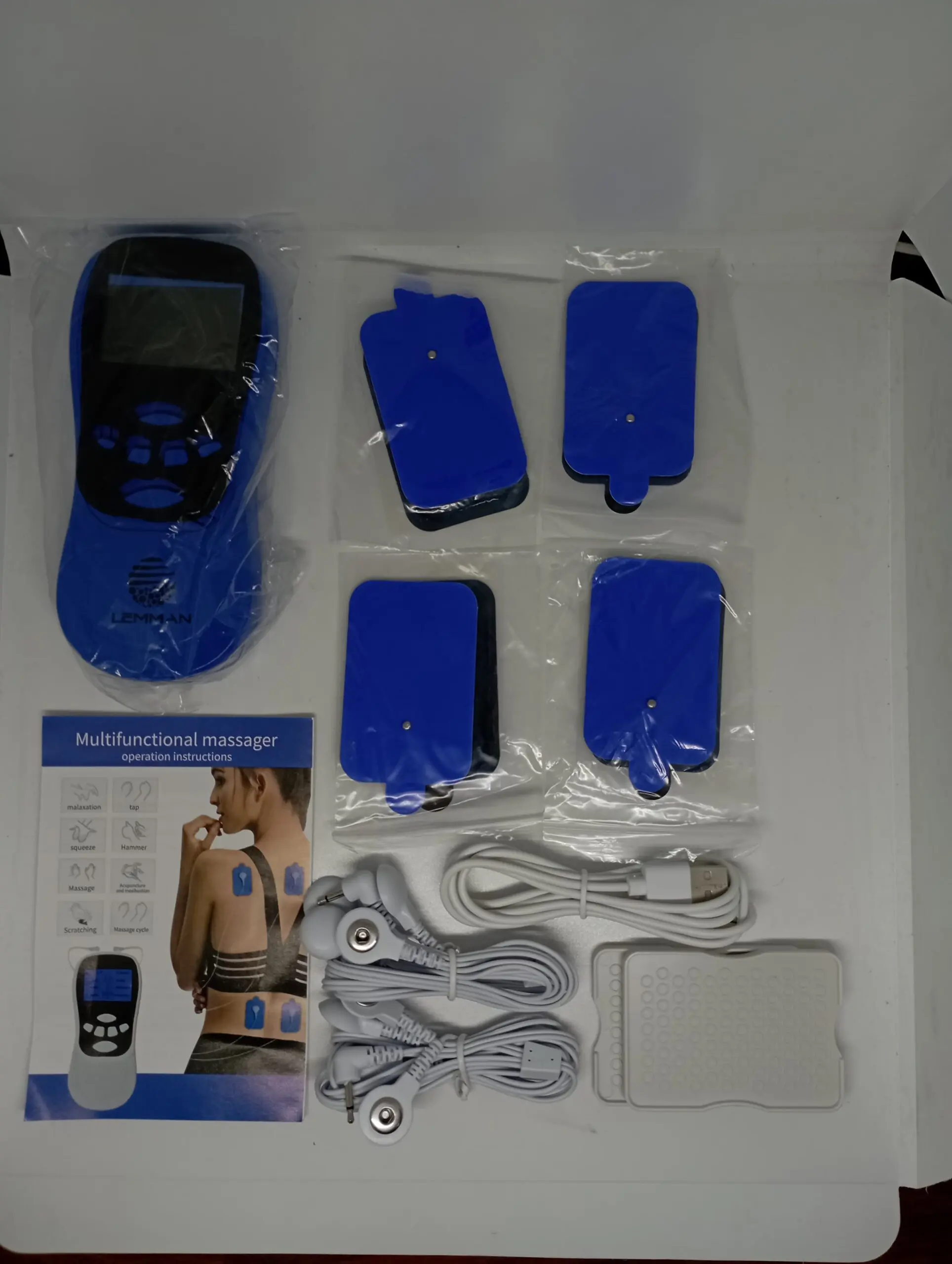 Kit de masajeador TENS digital azul con pantalla, cuatro parches, cables, cable USB, manual y almohadilla blanca dispuestos en su caja blanca.